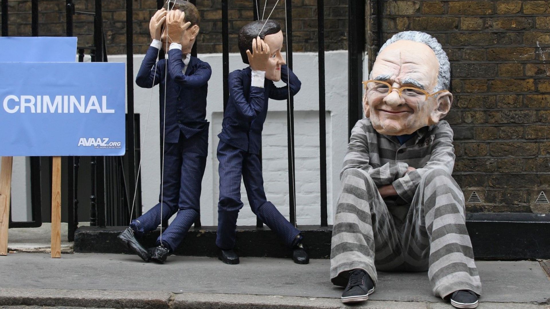 Протестиращ с маска на Рупърт Мърдок демонстрира с кукли на политическите лидери на консерваторите и лейбъристите - Дейвид Камерън и Ник Клег, пред апартамента на изпълнителния директор на News Corp на 13 юли 2011 г. покрай скандала с хакването на телефона на тийнейджър от News of the World